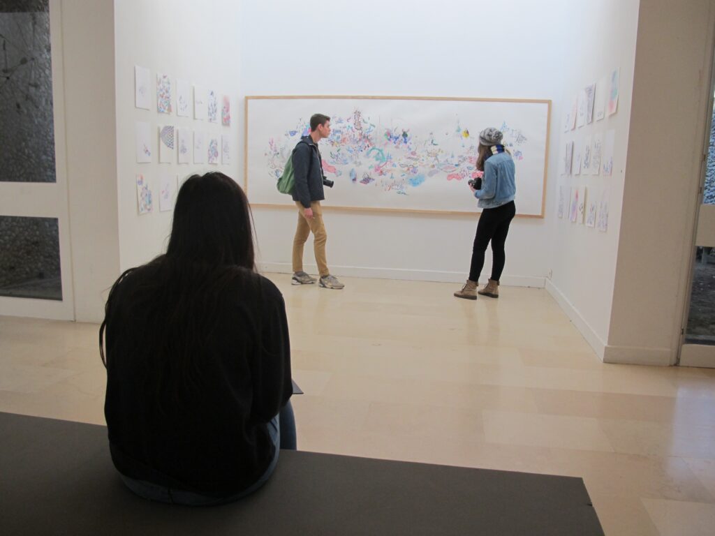 1 visiteurs sont debout devant une oeuvre alors qu'une personne les regarde assise sur un banc