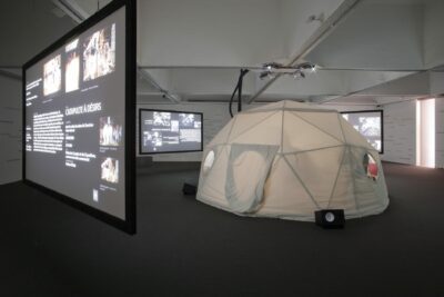 Sas d'entrée, 4 vidéoprojecteurs, 4 écrans de retroprojection, enceintes et caissons de basses amplifiés, moquette, sièges, lettrages adhésifs collés aux murs, bureau central (armature métallique, toile), ordinateurs, mobilier, éclairage, climatisation), installation au format de la salle le bureau de Cédric Moris Kelly et de Christine Bavière a été installé pour 4 mois sous une tente au centre de la salle