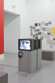 Une télévision catodique posé sur un socle avec en fond des papiers disposés sur un mur